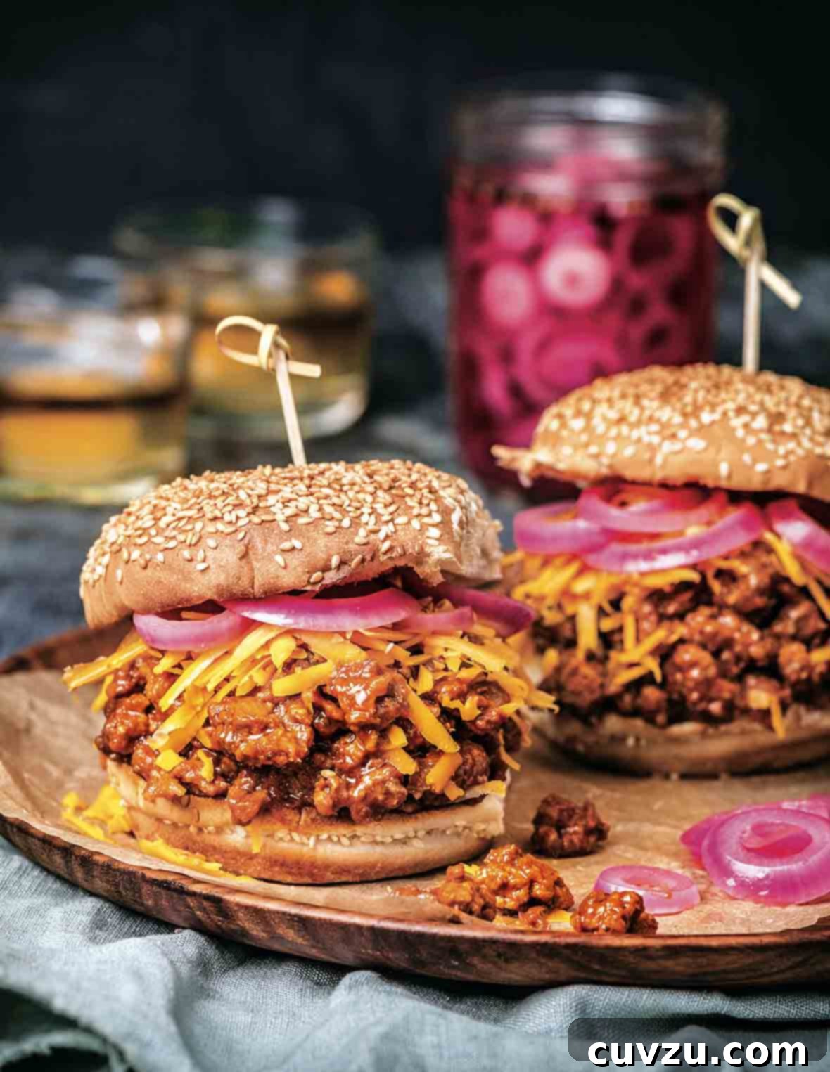 Two bourbon sloppy joes on sesame buns, topped with shredded Cheddar cheese, and pickled red onion, ready to eat.