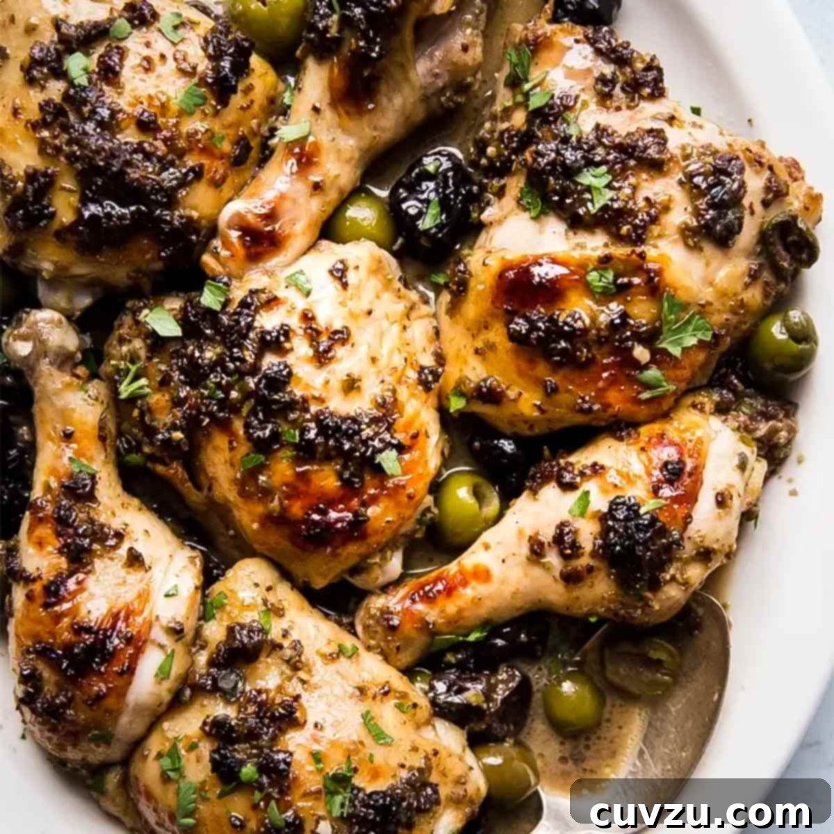 Pieces of chicken Marbella, topped with plums and herbs, with olives scattered around, on a white platter.