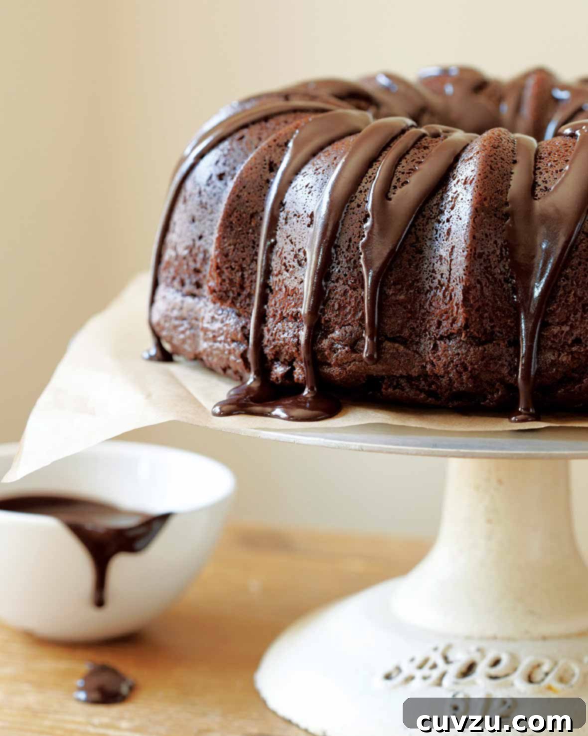 A chocolate glazed chocolate bundt cake sitting atop a white cake stand with a dish of glaze on the side
