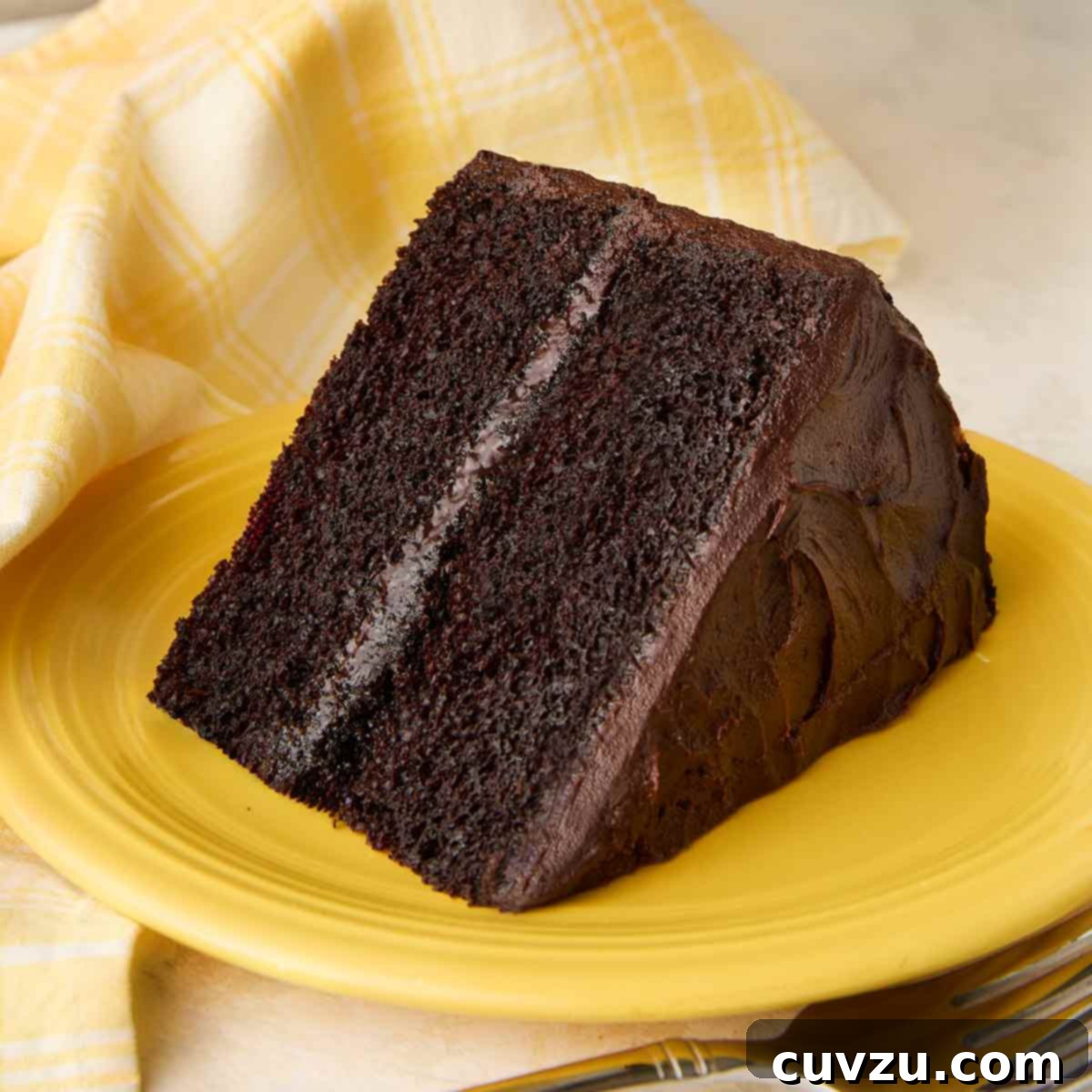 A slice of Hershey's chocolate cake with chocolate frosting on a yellow plate, with the remaining cake on a stand in the background.
