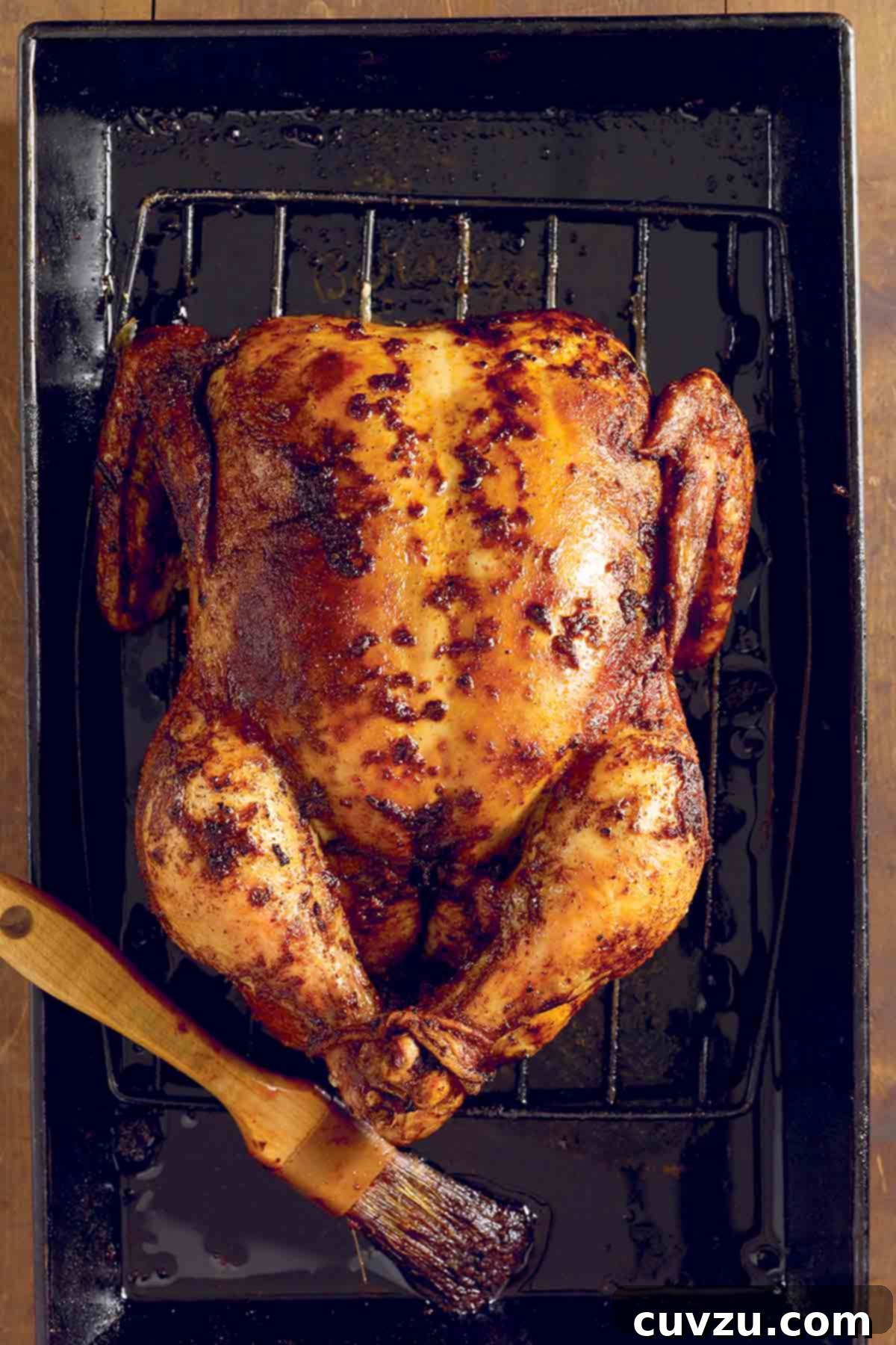 A whole, glistening spicy roast chicken resting on a rack inside a baking dish, with a basting brush lying beside it, tempting with its vibrant, fiery hue.