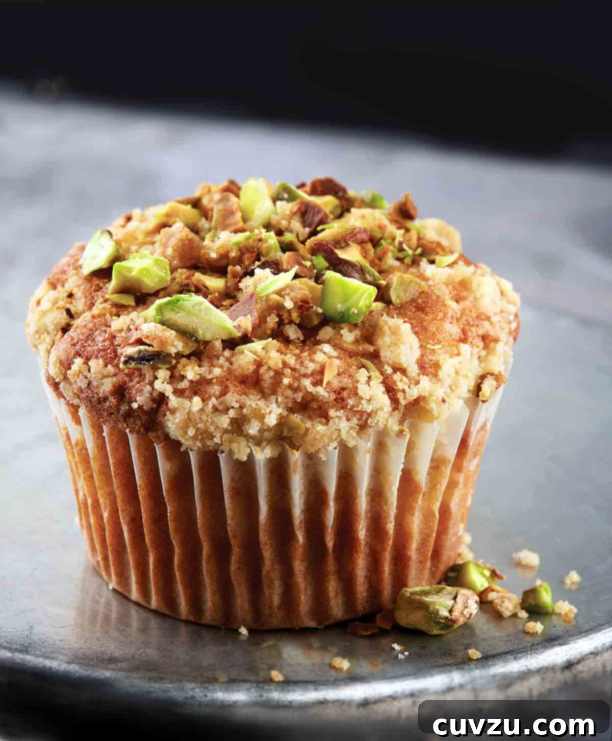 A single, perfectly domed apple spice muffin with a glistening streusel and chopped pistachios, resting invitingly on an inverted metal cake tin.