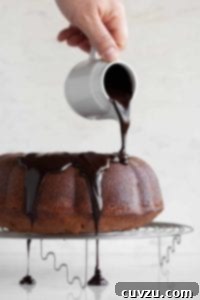 A person pouring a rich chocolate ganache glaze over a cooling orange chocolate chunk Bundt cake.