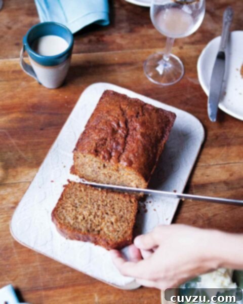 A person cutting a slice from a loaf of whole wheat banana bread on a white cutting board.