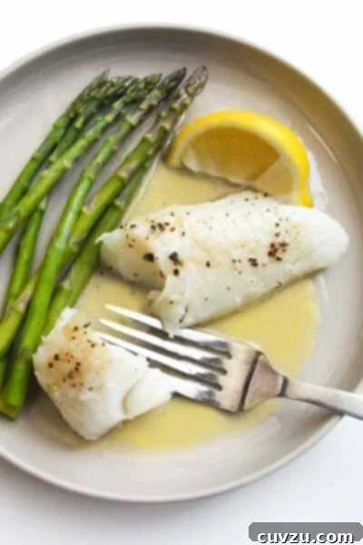 A piece of baked fish on a plate with butter sauce, a lemon wedge, and some asparagus spears.