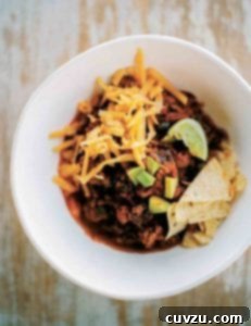 White bowl of beef chili with grated cheese, tortilla chips, and lime wedges on wood