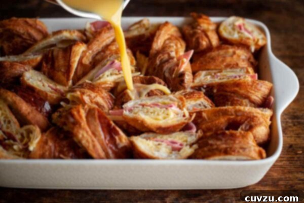 A generous stream of egg custard being poured over the arranged ham and cheese croissants in a baking dish, preparing the casserole for its overnight chill.