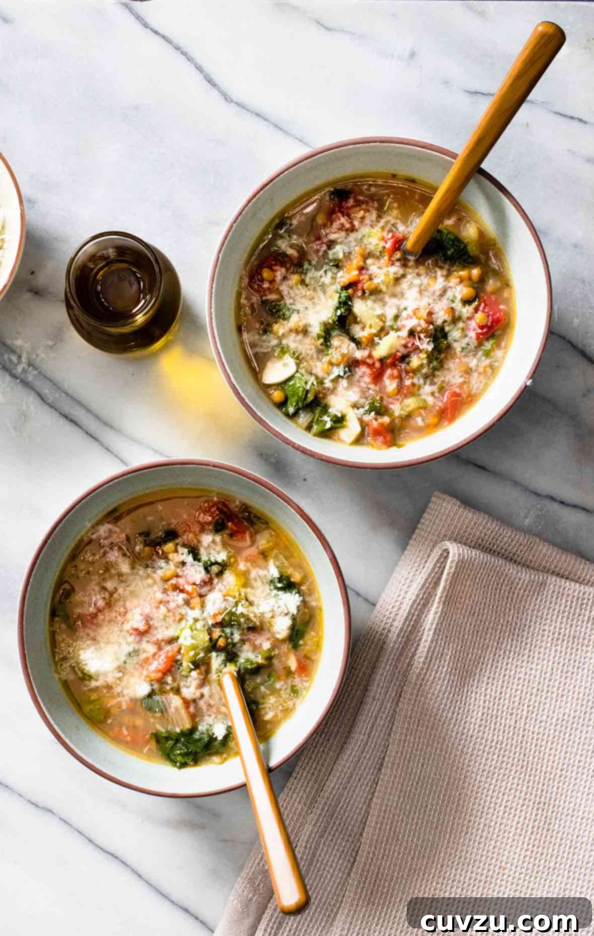 Two pristine white bowls brimming with hearty lentil and escarole soup, with a glass jar of olive oil resting nearby.