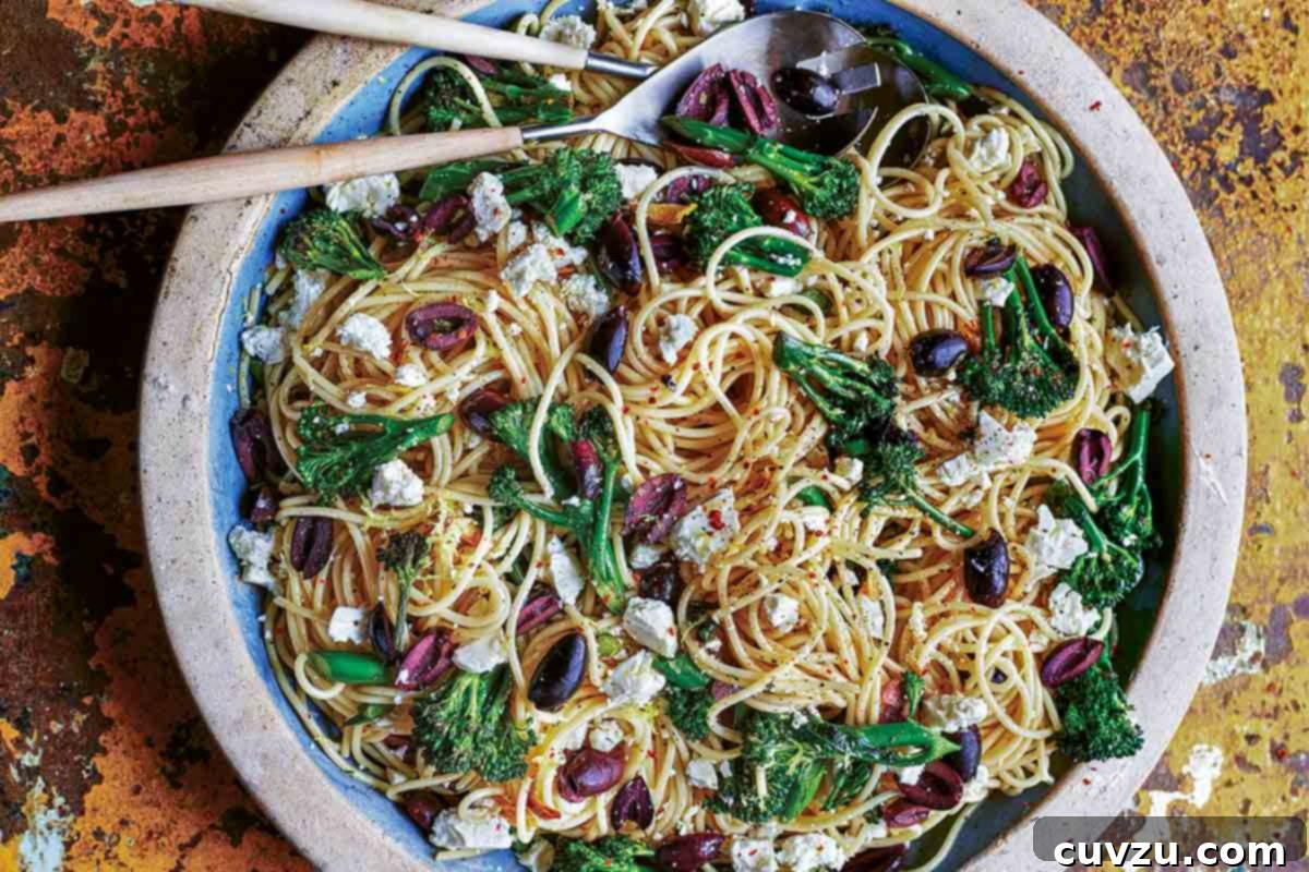 A vibrant plate of spaghetti tossed with briny olives, tender broccoli florets, and crumbled feta cheese, with two serving spoons nestled in the pasta.