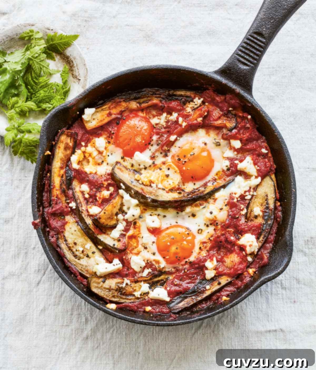 A rustic cast-iron skillet filled with flavorful eggplant shakshuka, featuring perfectly poached eggs nestled in a rich tomato and eggplant sauce, generously topped with crumbled feta cheese.
