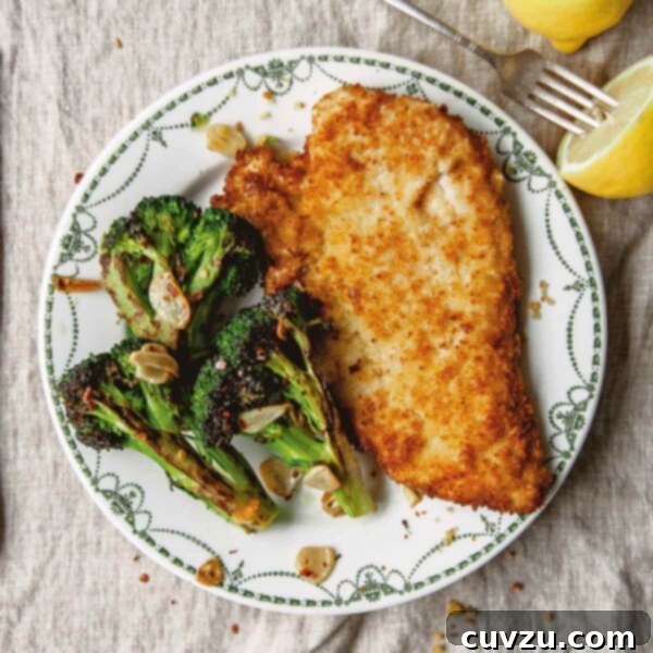 A piece of bread crumb coated chicken on a patterned plate with broccoli and a fork and two lemon halves on the side.
