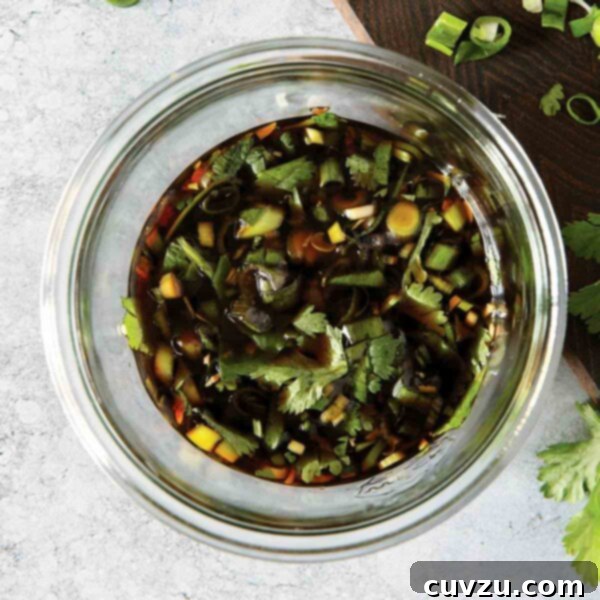 Ultimate Dumpling Dipping Sauce 3 A glass bowl filled with dumpling dipping sauce with pieces of scallion and cilantro floating in it and scattered around the jar, highlighting its fresh ingredients.