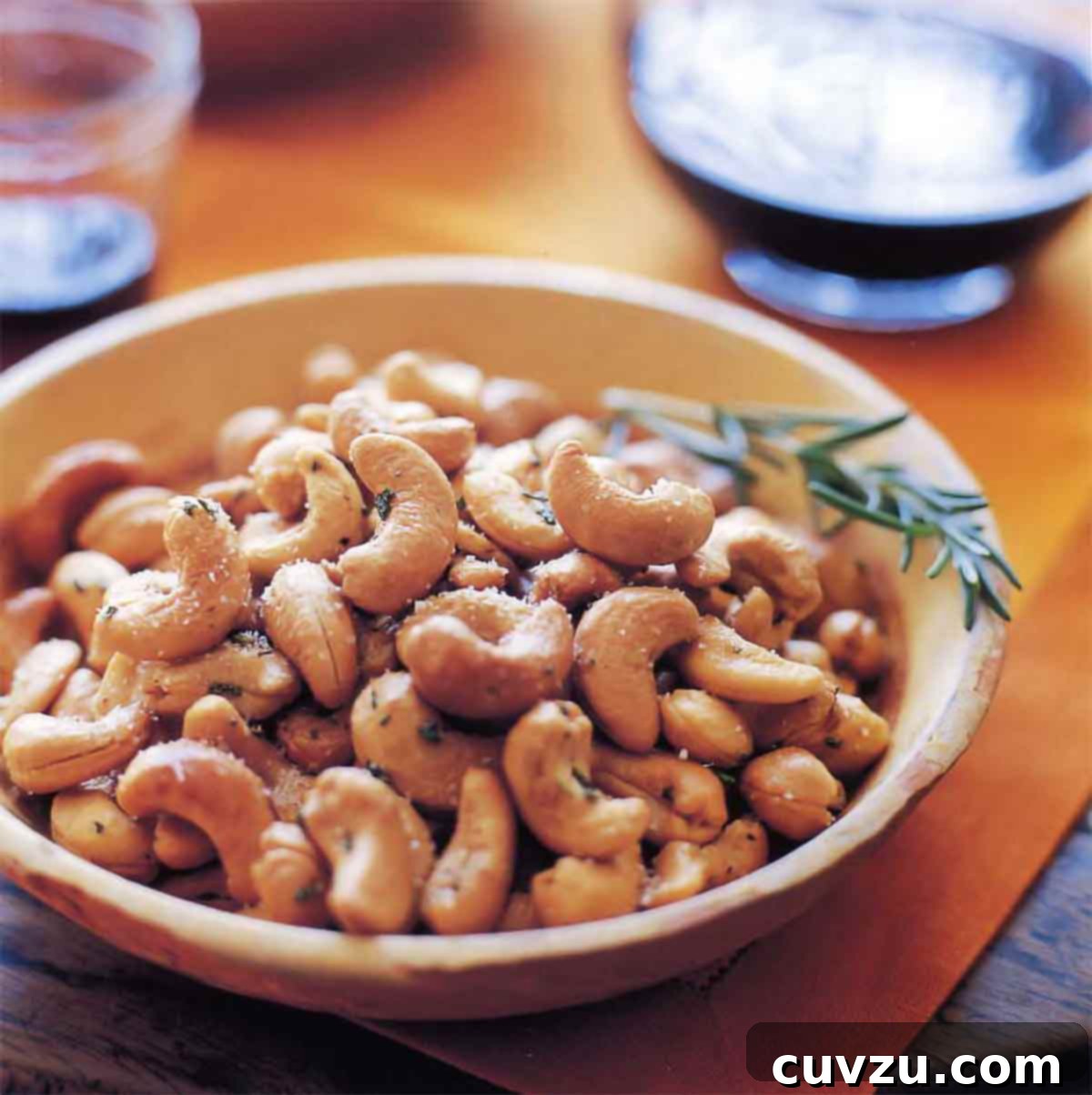A bowl filled with Ina Garten's rosemary cashews with a sprig of rosemary on the side, a perfect Barefoot Contessa snack.