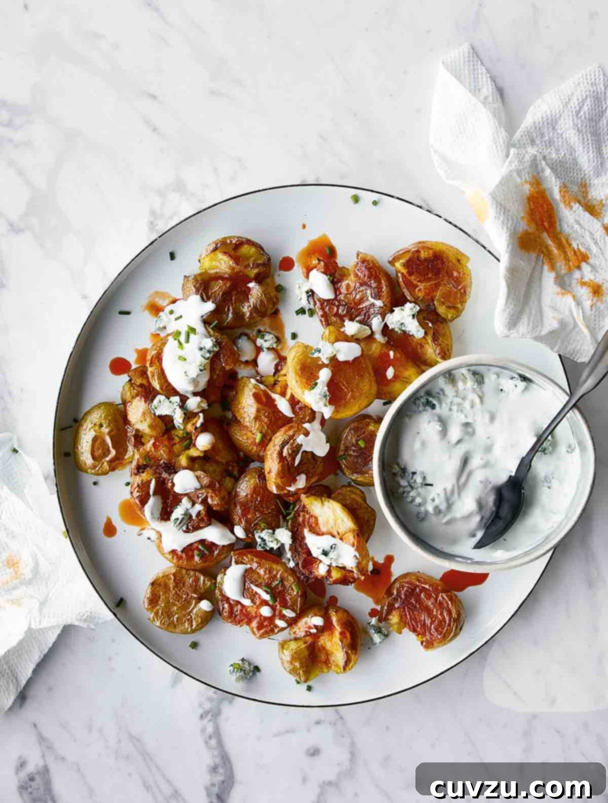 A white plate filled with crispy smashed buffalo potatoes with a bowl of blue cheese sauce on the side and a spoon resting inside.