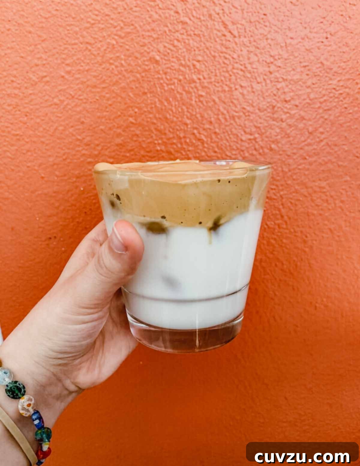 Whipped coffee in a clear glass, being held in front of an orange wall, showcasing its beautiful layers.