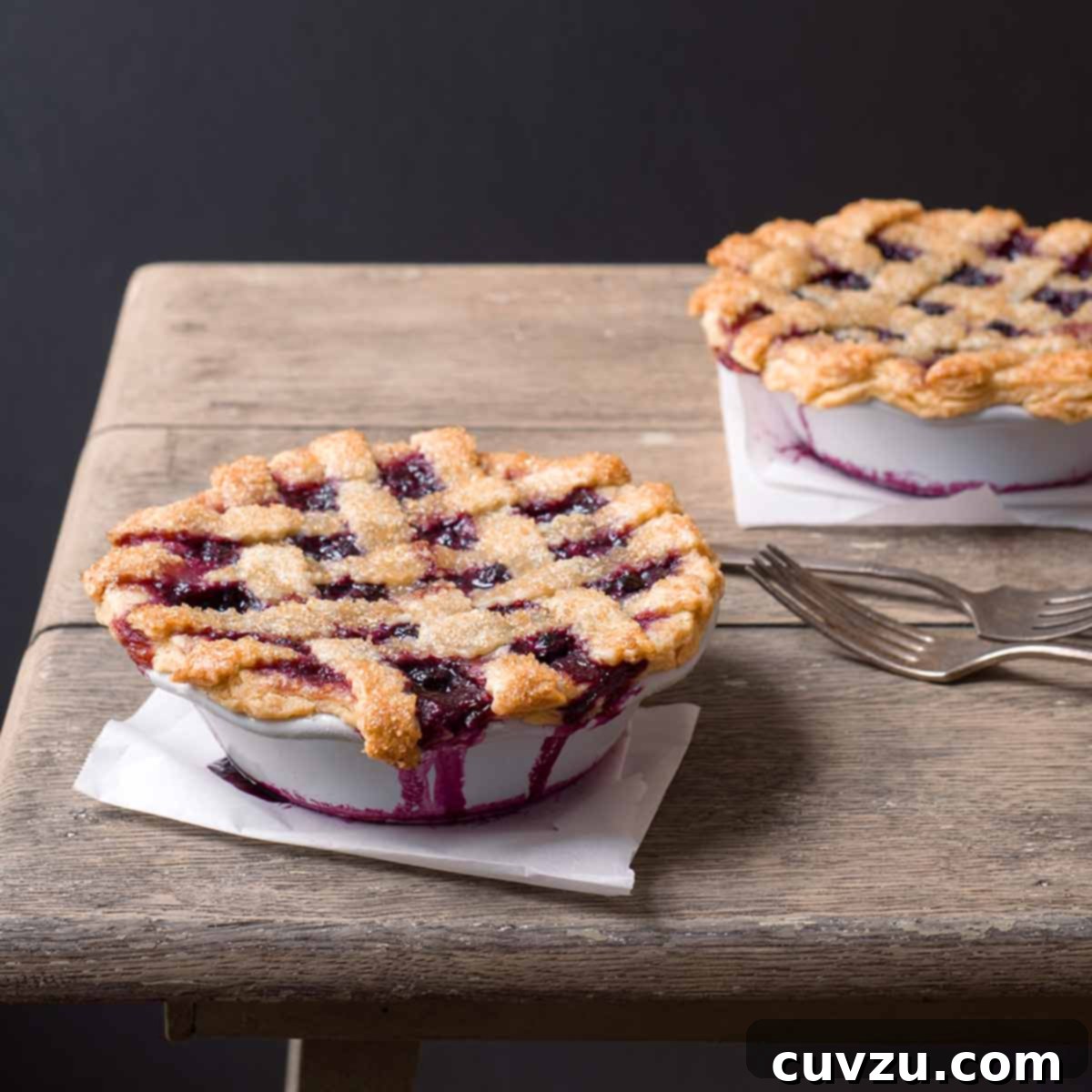 Two individual blueberry pies on a wooden table with forks beside them.