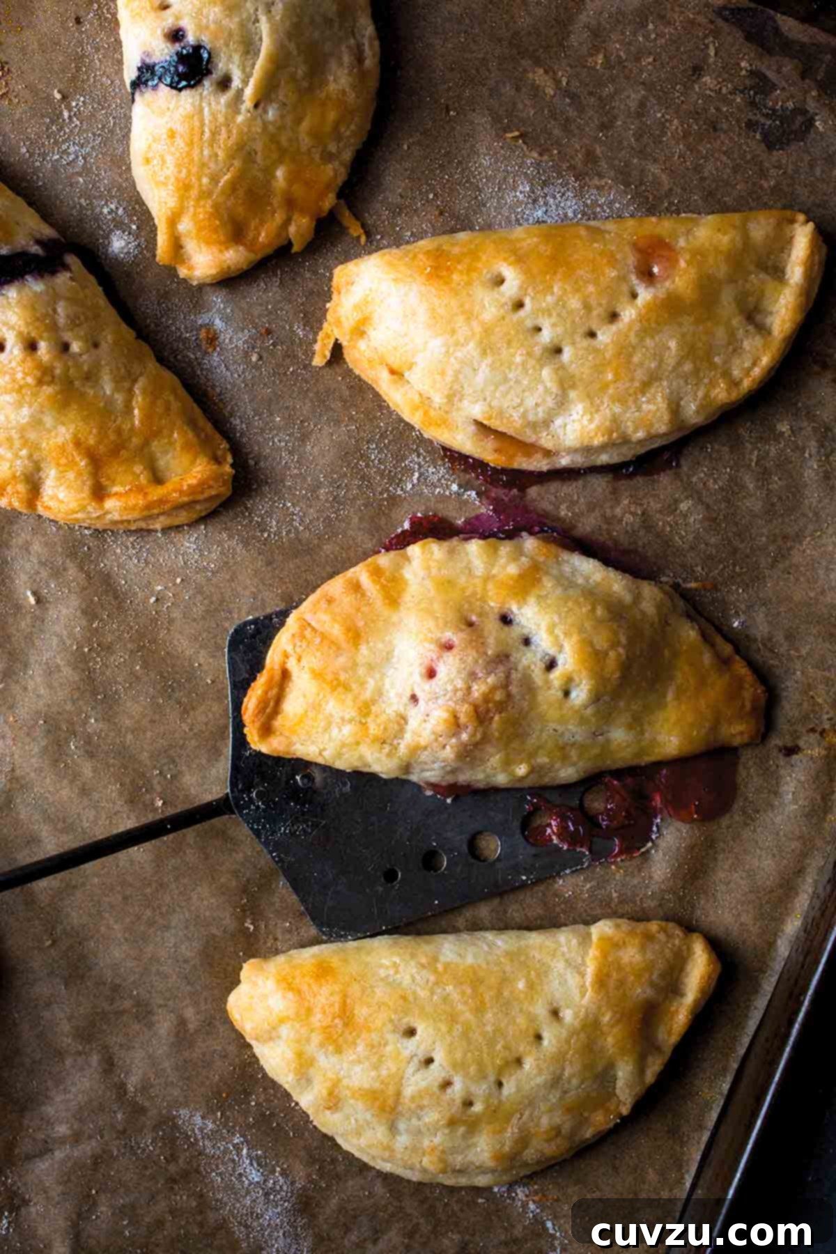 Five hand pies on a parchment-lined baking sheet with a spatula lifting one off.