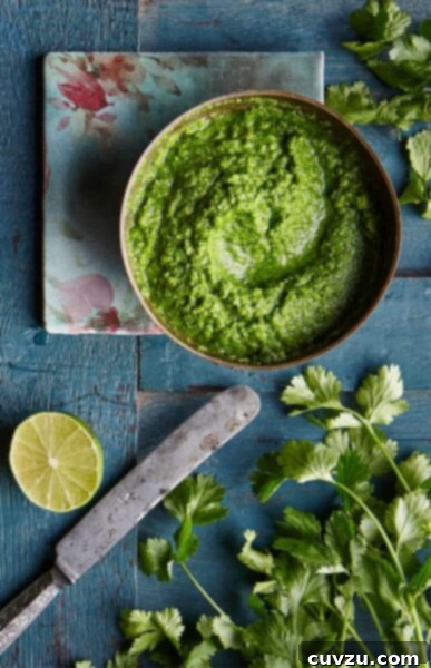 A bowl of bright green cilantro peanut chutney on a blood wooden table with a flower napkin, cilantro leaves, half a lime, and a knife.
