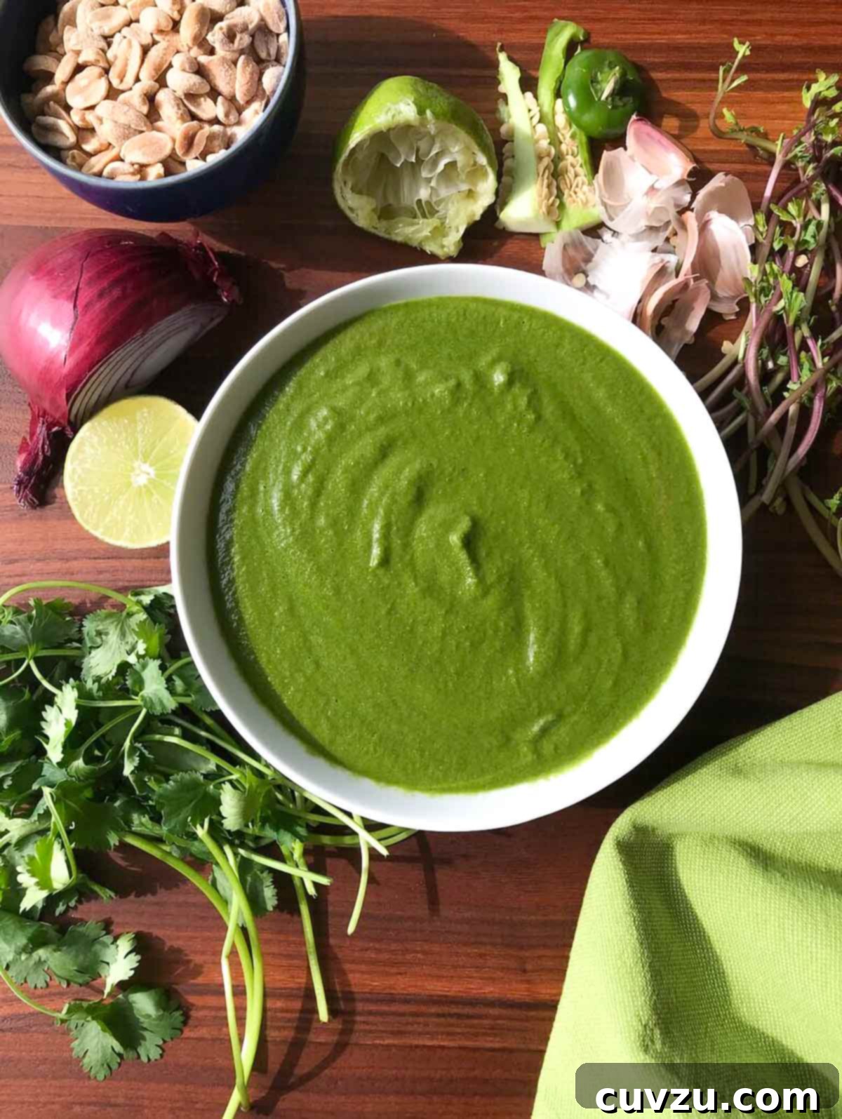 A white bowl of bright green cilantro peanut chutney, artfully arranged on a wooden table alongside bowls of peanuts, fresh cilantro, lime wedges, garlic cloves, and onions, ready for blending.