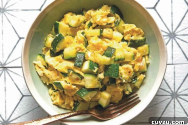A green bowl filled with scrambled eggs and green pieces of zucchini on a tile tabletop, with a fork.