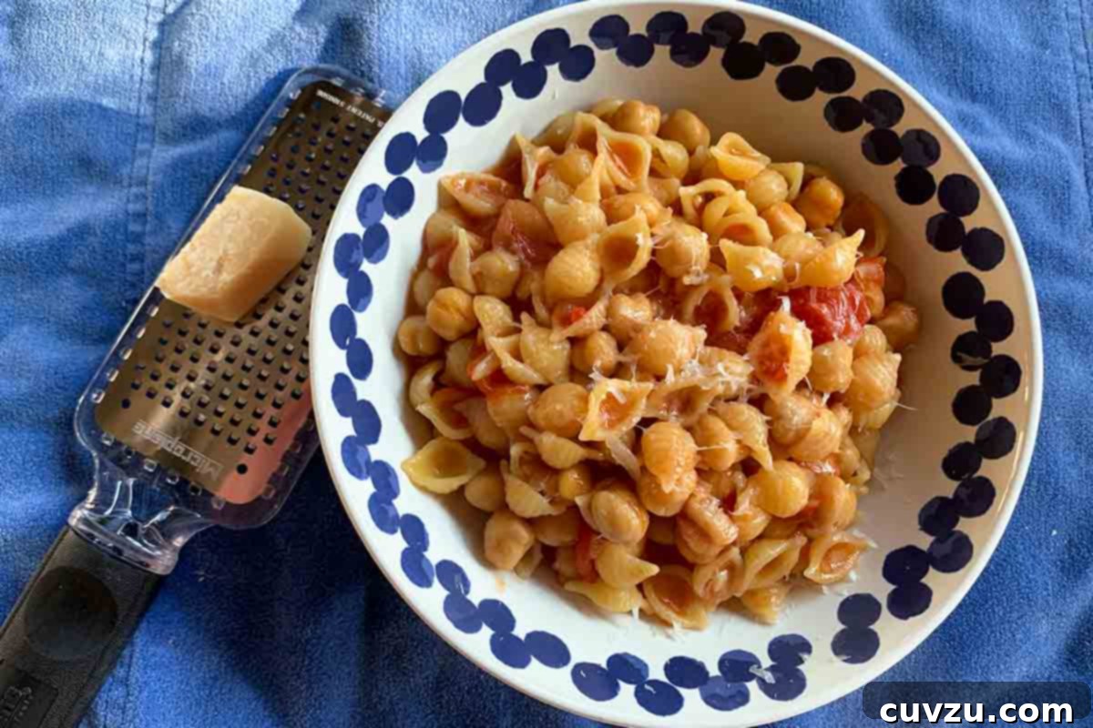 Rustic Chickpea Tomato Pasta 5 A blue and white bowl filled with pasta shells, tomatoes, chickpeas, and shavings of Parmesan, beside a grater and a chunk of Parmesan, highlighting the rustic appeal of the dish.