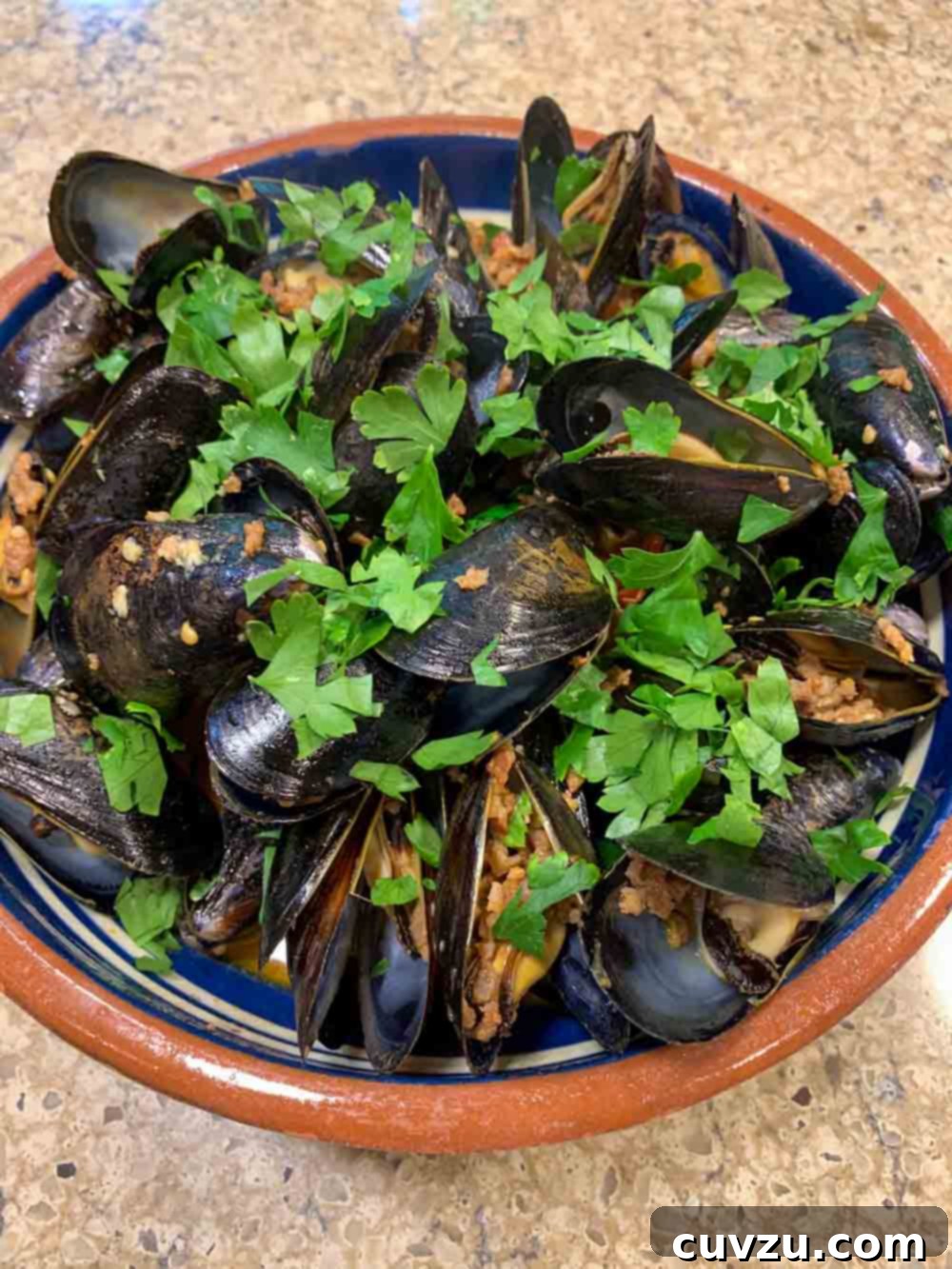 A rustic blue and terracotta bowl overflowing with plump, steamed mussels, vibrant cherry tomatoes, and chunky pieces of chorizo, all generously garnished with fresh green parsley. The rich broth pools invitingly at the bottom.