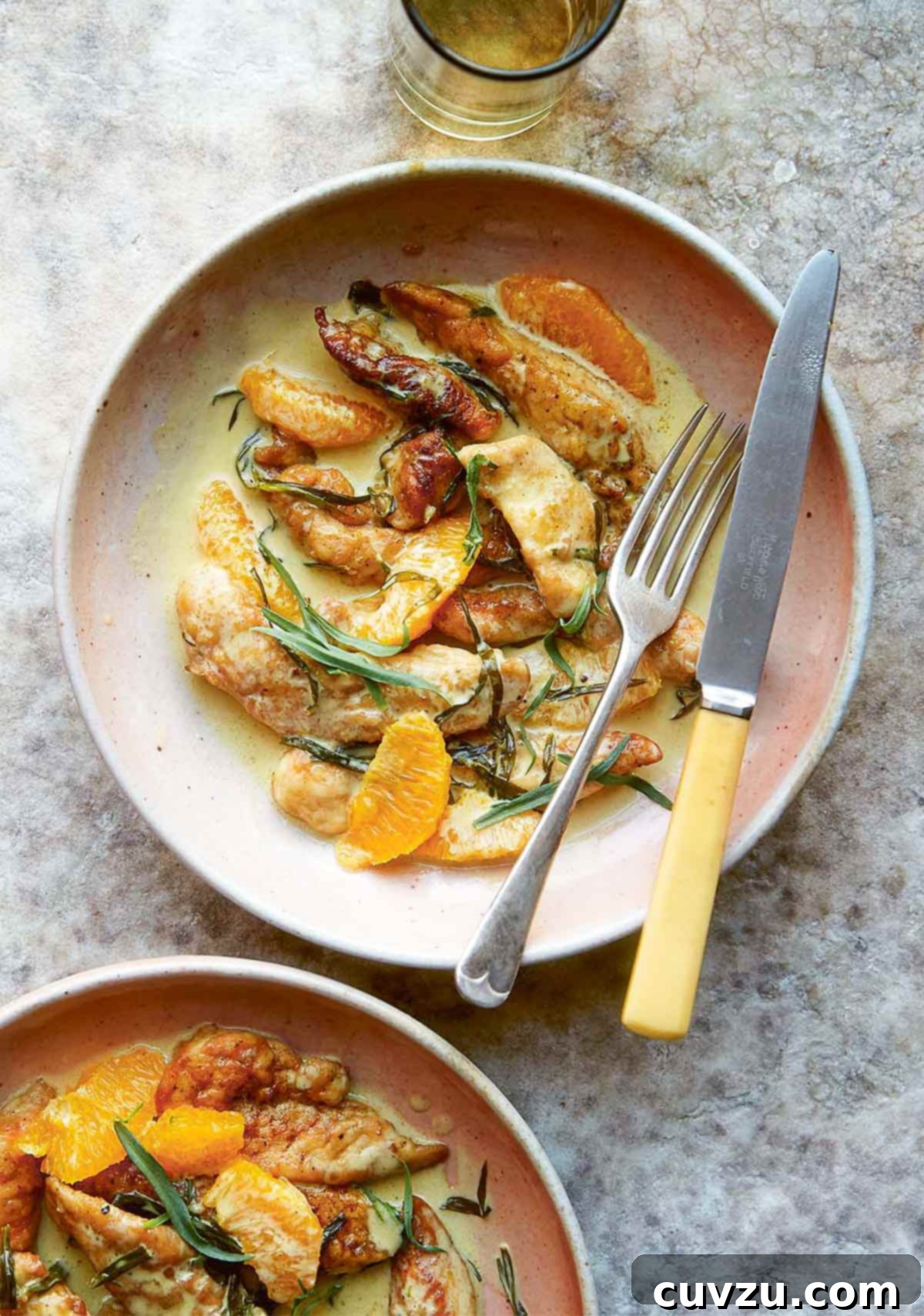 Two bowls of chicken tenders in a rich cream sauce, adorned with vibrant orange slices and delicate chiffonaded tarragon leaves, presented alongside a knife and fork.