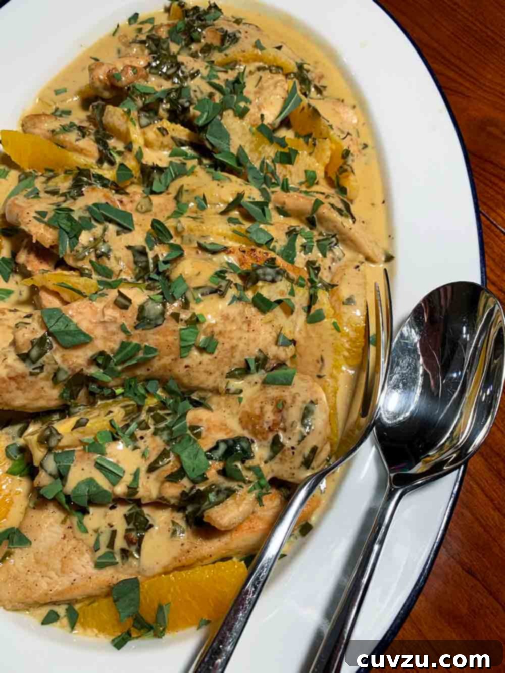 An oval white platter covered with strips of chicken breast, orange slices, cream sauce, and tarragon, with serving utensils.