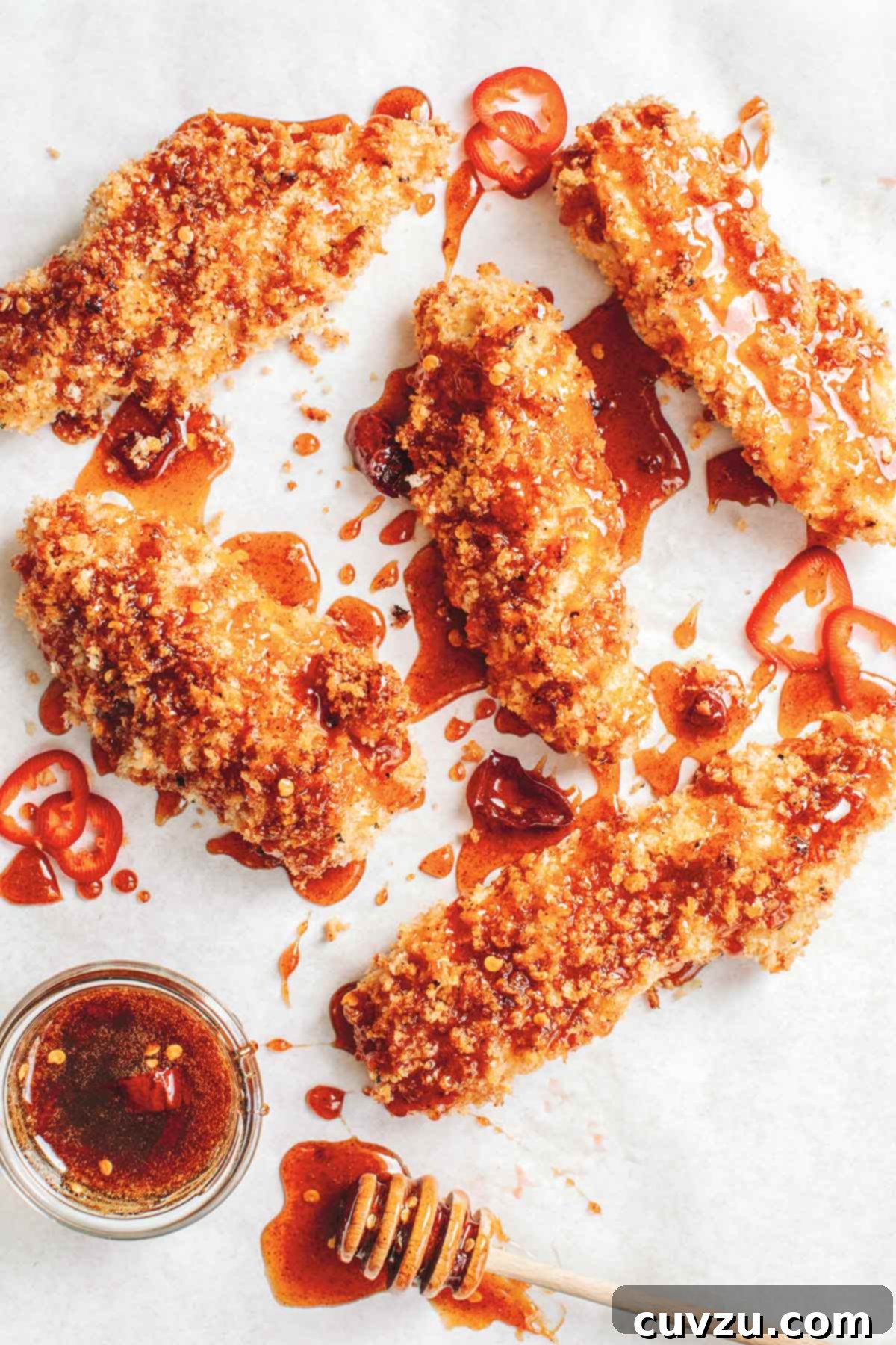 5 panko breaded chicken tenders drizzled with deep red hot sauce and chile slices, with a bowl of more spicy honey and a honey dipper.