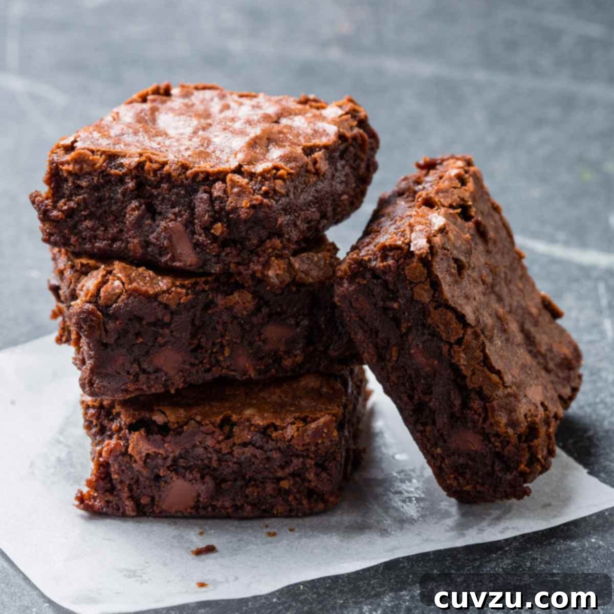 Three chewy chocolate brownies stacked on top of each other with another leaning on the stack on a piece of parchment.