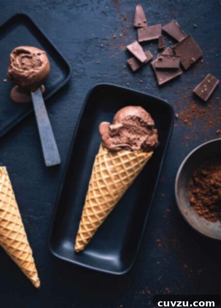 Two rectangular plates with scoops of dark chocolate gelato, one on a scoop and one in a cone, surrounded by squares of dark chocolate and a bowl of cocoa powder. This rich, smooth, and extra chocolatey gelato is perfect for discerning palates.