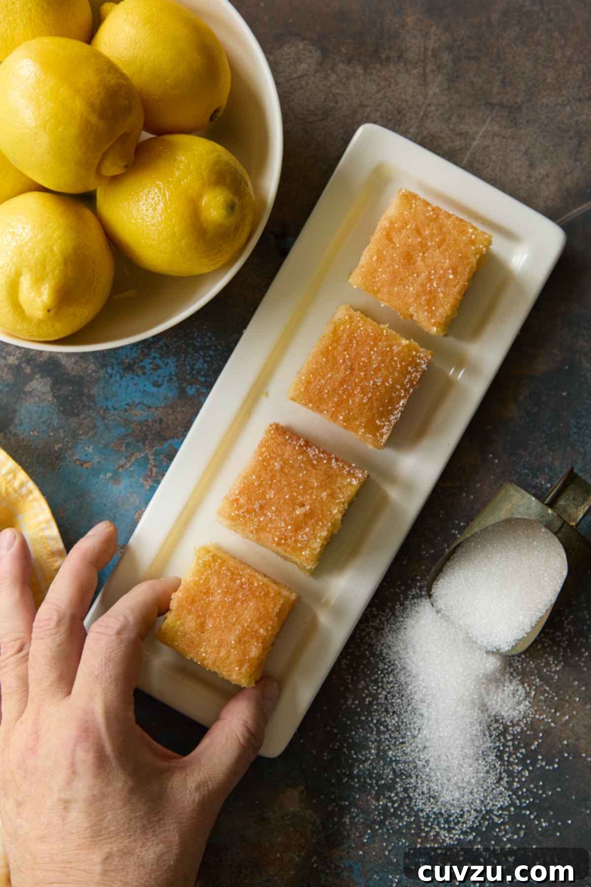 Zesty Lemon Loaf 15 Overhead photo of lemon drizzle cake slices on a white tray, fresh lemons in a bowl, and a hand reaching for a piece next to spilled granulated sugar.
