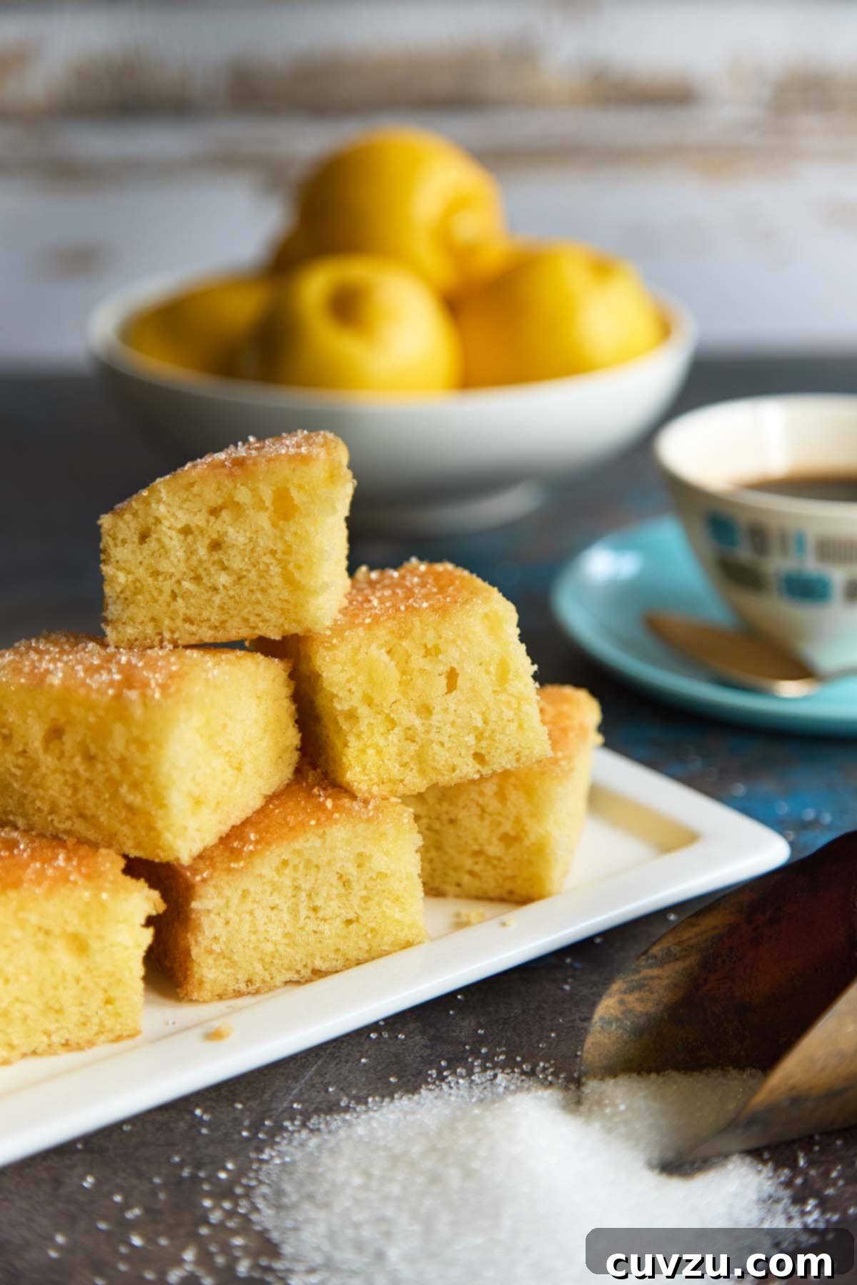 Zesty Lemon Loaf 16 Stack of moist lemon drizzle cake squares on a white platter, sprinkled with sugar, next to a bowl of lemons and a cup of coffee.