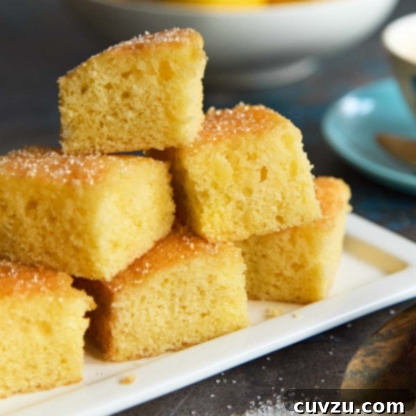 Zesty Lemon Loaf 17 Moist lemon drizzle cake squares stacked on a white plate with a sugar crust, next to a cup of coffee and a bowl of lemons.