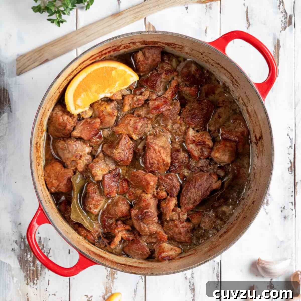 A vibrant red Dutch oven filled with succulent braised pork cubes, adorned with an orange wedge.