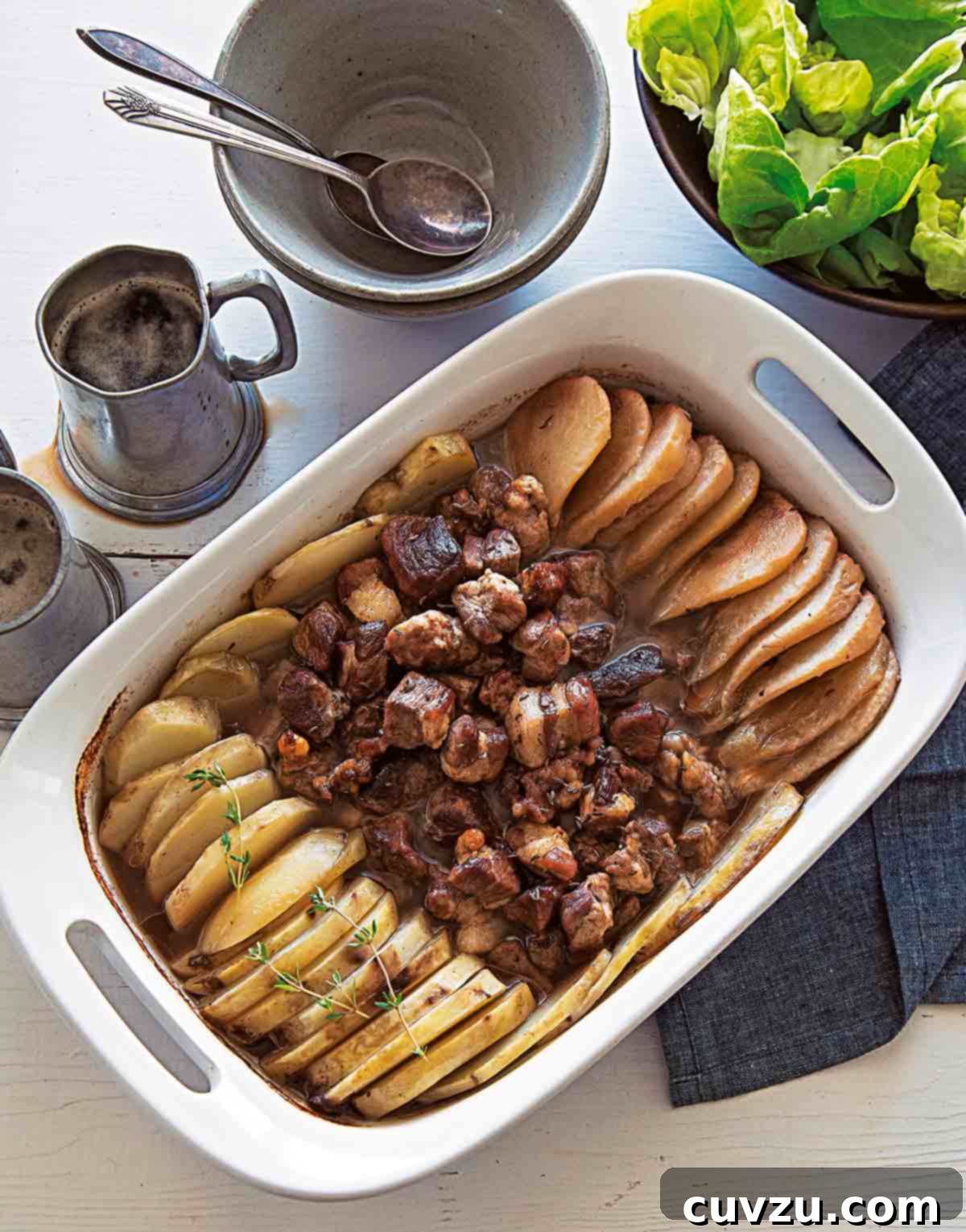 A white casserole dish brimming with a delectable roast pork, potatoes, and pears, served alongside a fresh salad.