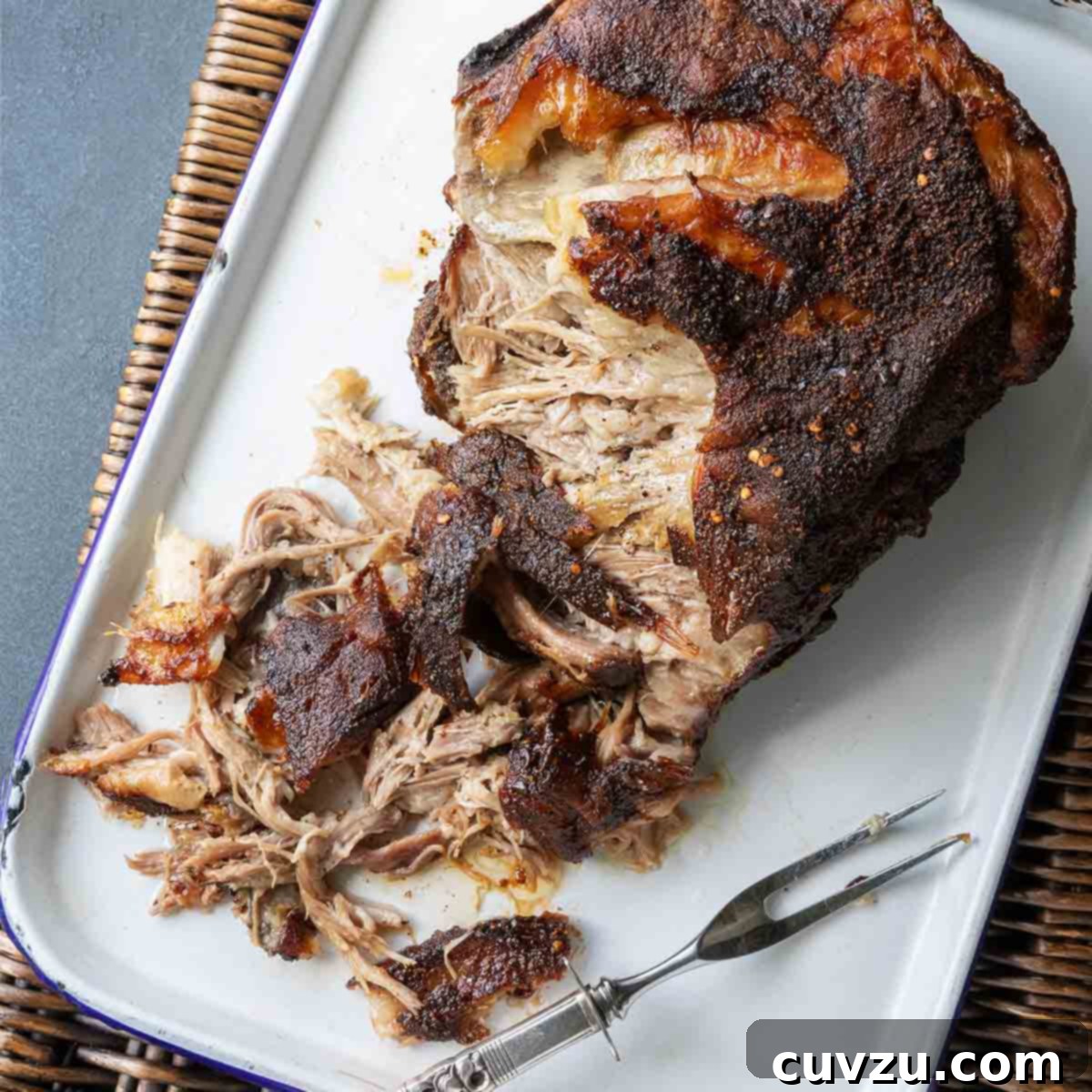 A partially shredded roast pork butt in a roasting pan with a fork nearby, showcasing its tender texture.