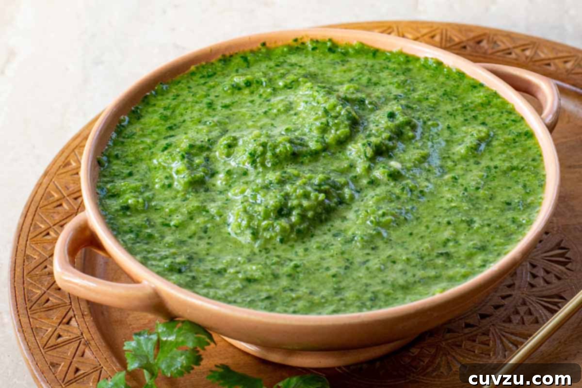 The Yemeni Green Flame 2 A bowl of vibrant green Schug, garnished with fresh cilantro, resting on a rustic wooden plate with a spoon beside it.