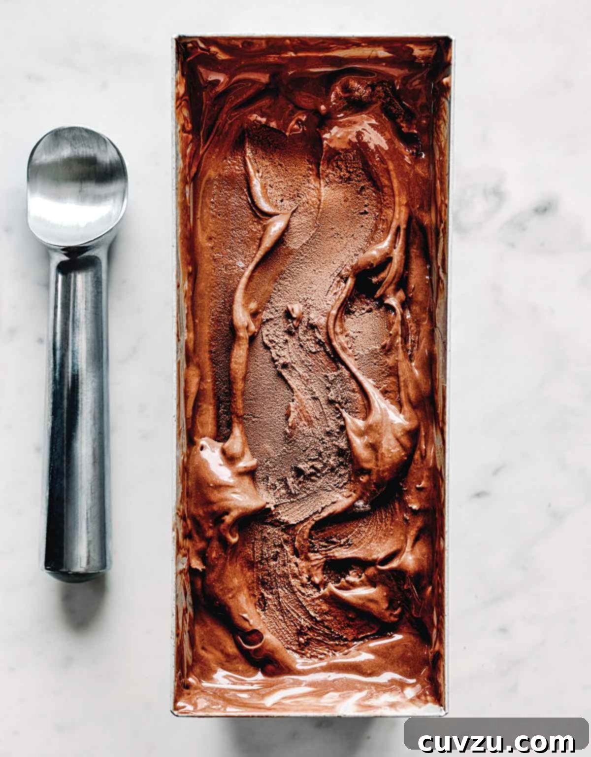 Dark chocolate tahini ice cream in a metal ice cream tin with a swirl on top, with a metal ice cream scoop laying beside it.