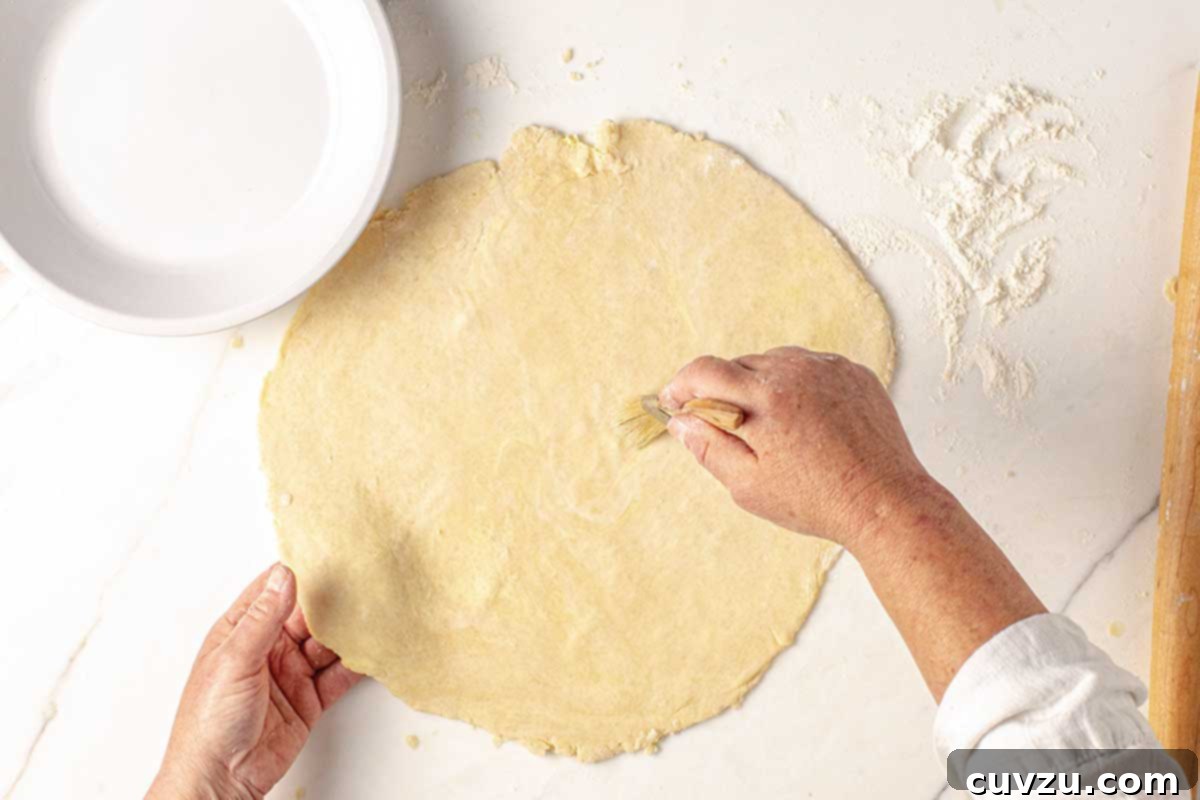 Roll Out a Flawless Pie Crust Every Time 6 A person using a pastry brush to remove excess flour from a rolled out pie crust, ensuring a smooth, clean surface.