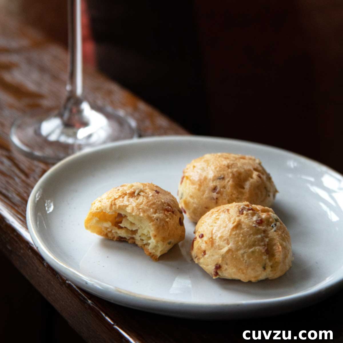 A white plate with three prosciutto-cheese gougeres with a glass of Prosecco in the background.