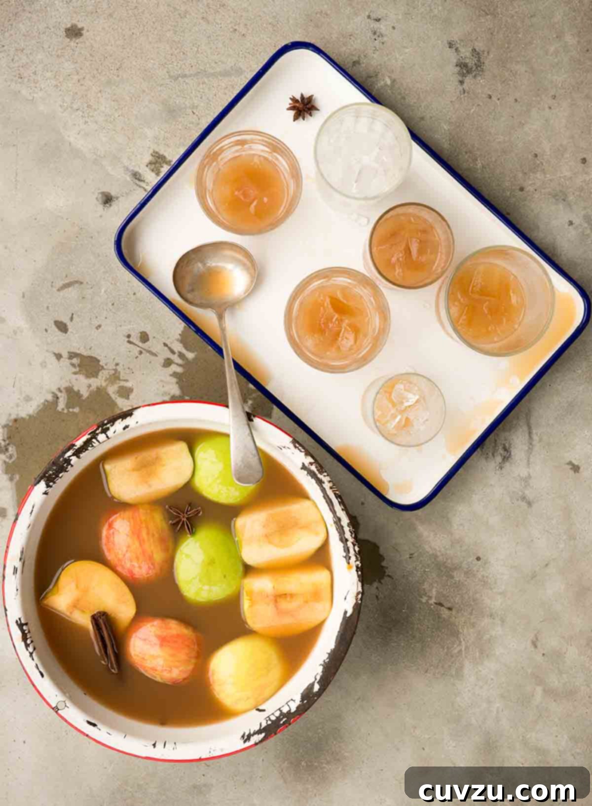 A chipped pot filled with apple pie moonshine, apple wedges and whole spices with a tray of glasses filled with the moonshine beside it.