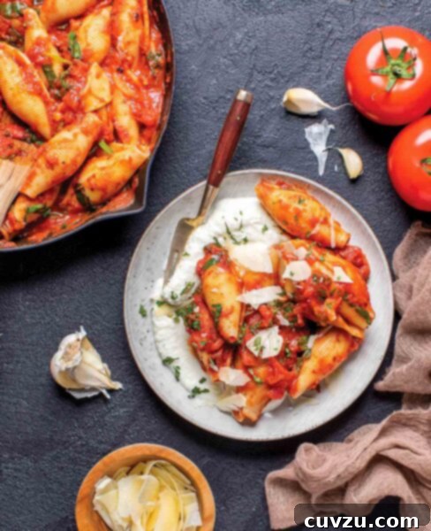 Shells with ricotta and fire-roasted tomato sauce on a white plate with a fork, beside a pan of shells, a clove of garlic, parmesan, and a tomato.