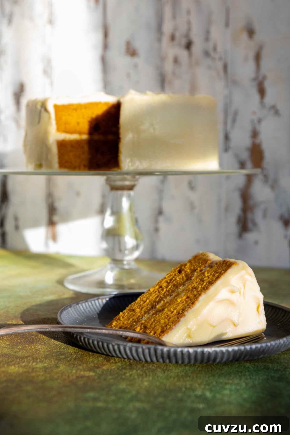 A slice of frosted pumpkin cake on a plate with the remaining cake on a stand in the background.