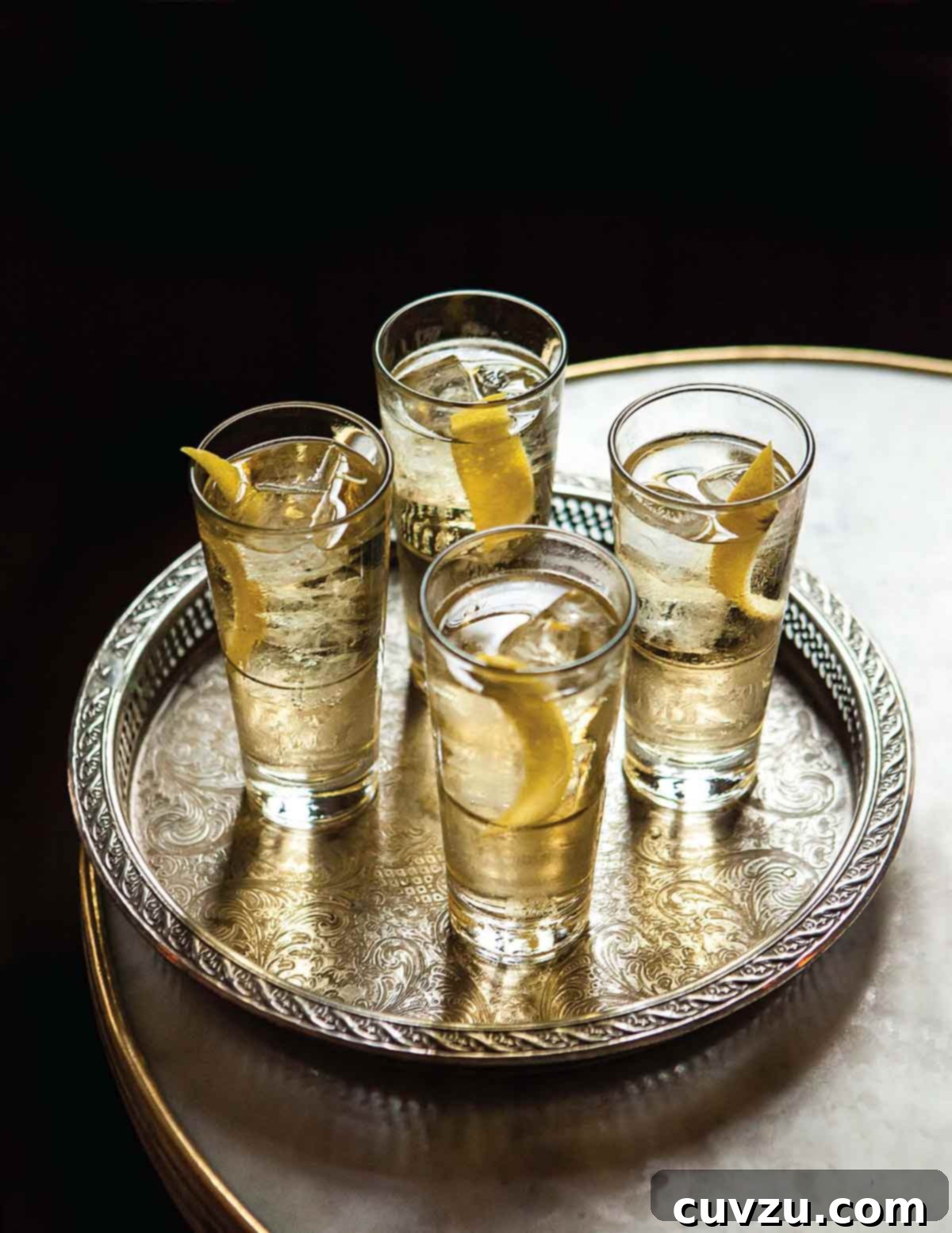 4 delicate tumblers filled with St. Germain Cocktail with a little ice and a lemon twist, sitting on a silver tray on a marble table.