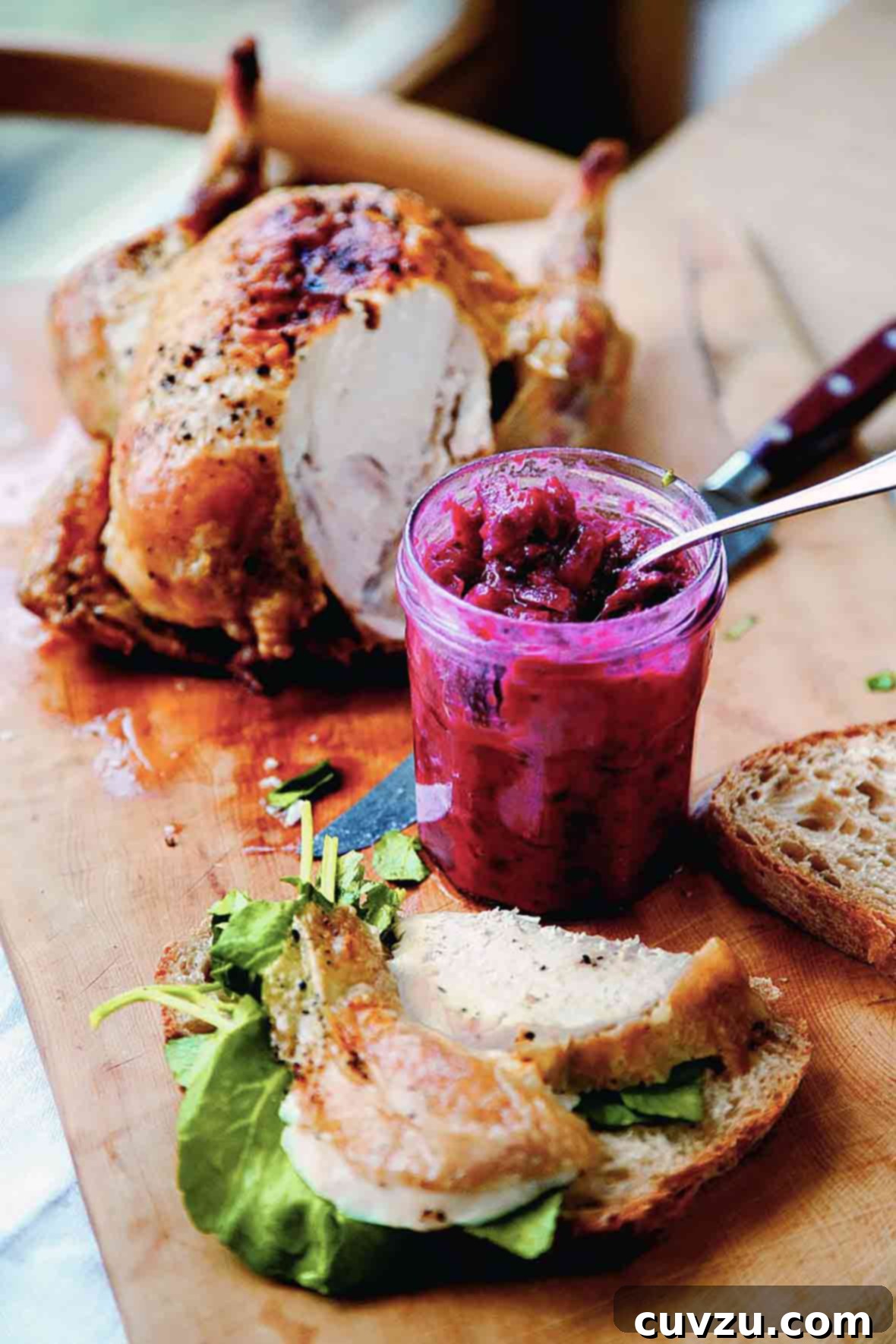 An open jar of cranberry chutney on a cutting board next to a partially carved chicken and a sandwich being assembled.