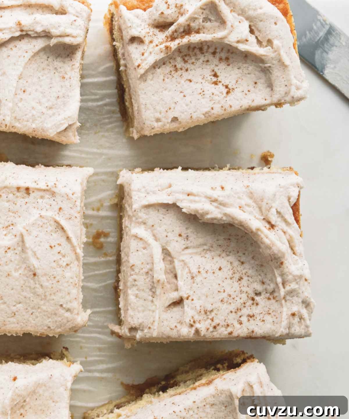 Golden Brown Butter Snickerdoodle Cake 2 Brown butter snickerdoodle cake cut into six pieces on a piece of parchment paper, with a serving knife nearby. Each slice reveals a beautiful cinnamon swirl and a generous layer of creamy frosting.