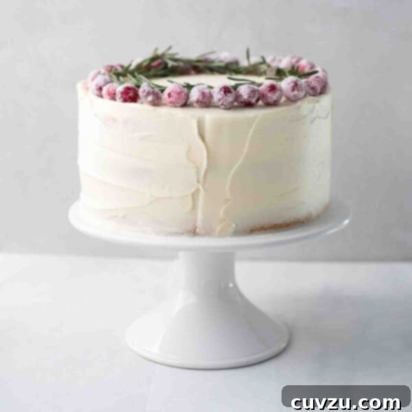 Velvet White Chocolate Christmas Cake 4 A whole frosted white Christmas cake topped with sugared cranberries and rosemary sprigs on a white cake stand.