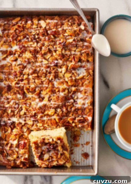 Make-ahead cinnamon coffee cake in a large rectangular pan, with two squares cut out, beside two cups of coffee and a bowl of drizzle with a spoon.