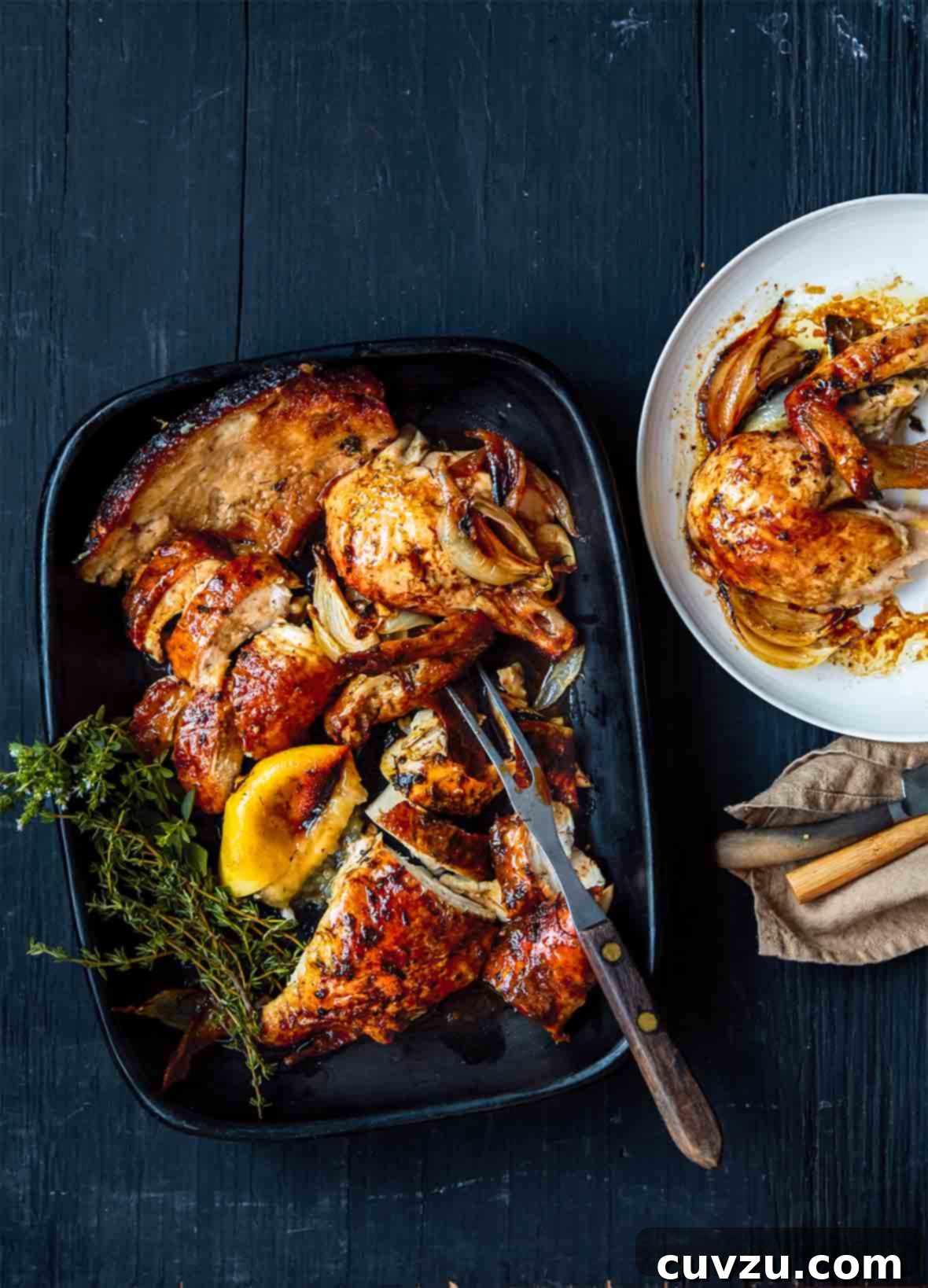 A delectable golden-brown herb butter chicken, roasted to perfection by Dorie Greenspan, elegantly arranged on a black platter alongside tender roasted onion, a bright lemon wedge, and fresh sprigs of thyme and oregano for garnish.